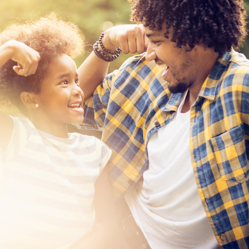 Father holds arm up in strong arm position as daughter smiles up at him.